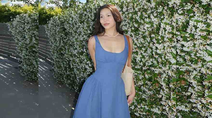 Chloe Kim in a blue sleeveless dress stands beside a white flowering hedge on a sunny day, with a beige shoulder bag.