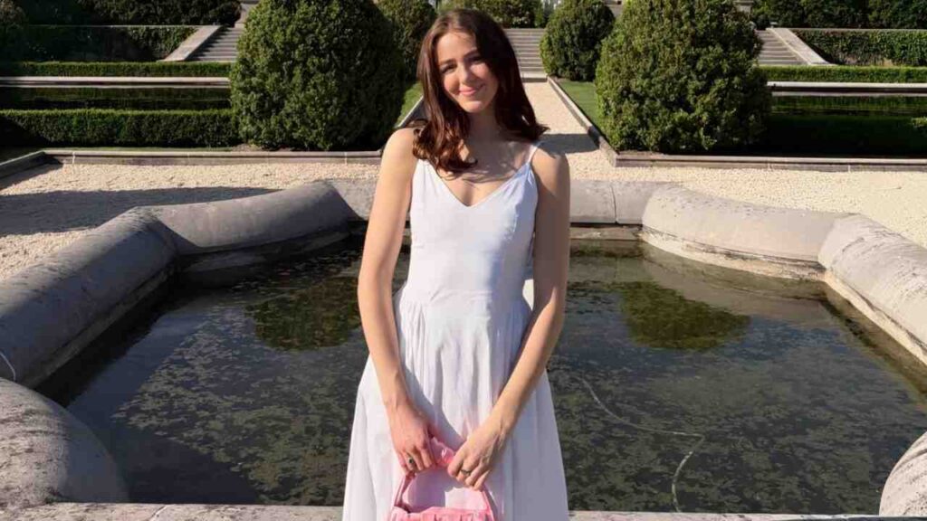 Chloe Lukasiak in a light lavender dress stands beside a stone fountain in a formal garden, holding a pink bag.
