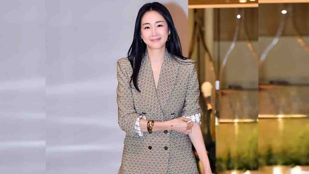 Choi Ji-woo in a beige geometric double-breasted blazer with rolled white cuffs and a gold bracelet, arms crossed against a gray and gold showroom backdrop.