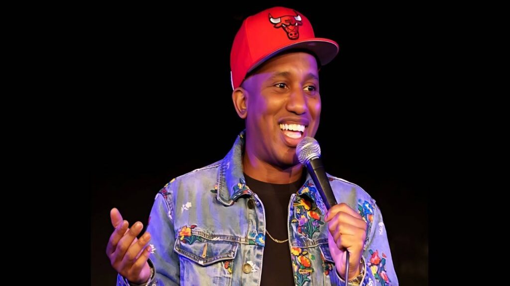 Chris Redd on stage holds a microphone, wearing a red Chicago Bulls cap and floral-embroidered denim jacket.