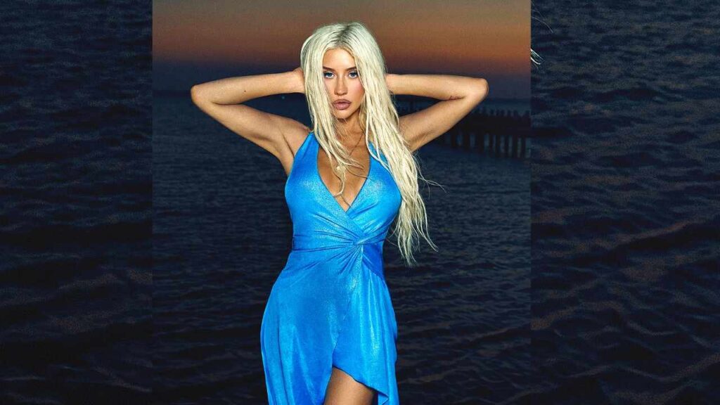 Christina Aguilera in a bright blue halter dress poses with arms behind her head by dark water at sunset, long hair, a pier visible