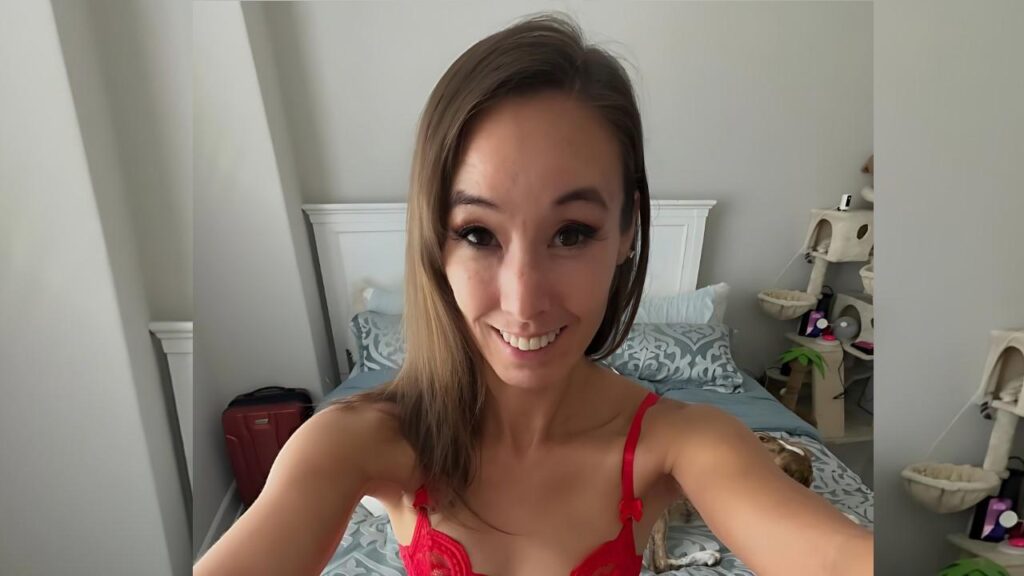 Christy Love in red lace bralette takes a selfie in a bright bedroom with a white headboard and blue-gray bedding.
