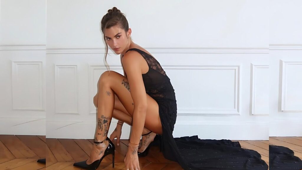 Clara Berry in a black lace dress crouches on a wooden floor in stilettos, paneled walls, train trailing.