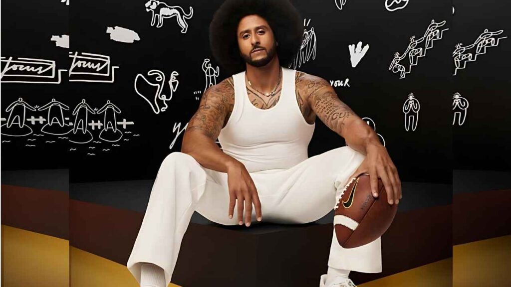 Colin Kaepernick sits on a black pedestal in a white tank, holding a football; tattooed arms, chalk drawings on a black wall.