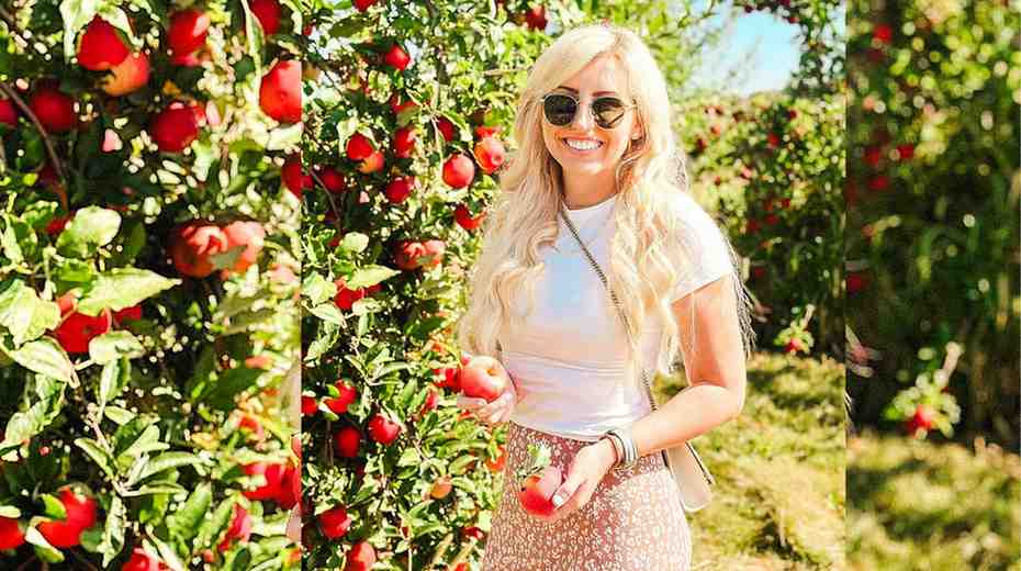 Courtney Force in a white top and floral skirt stands in an apple orchard, holding red apples.