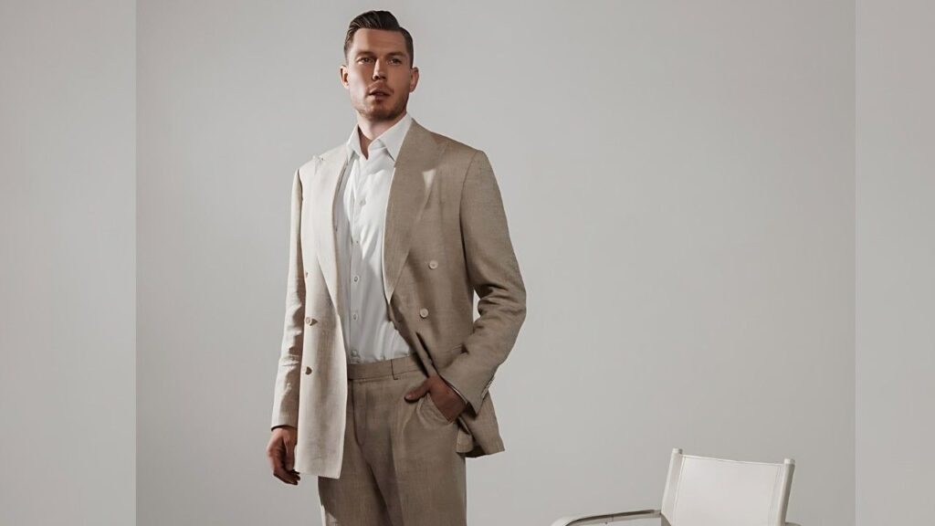 Dairis Bertāns in a light beige linen suit and white shirt stands in a minimalist studio, one hand in pocket, chair visible to the right.
