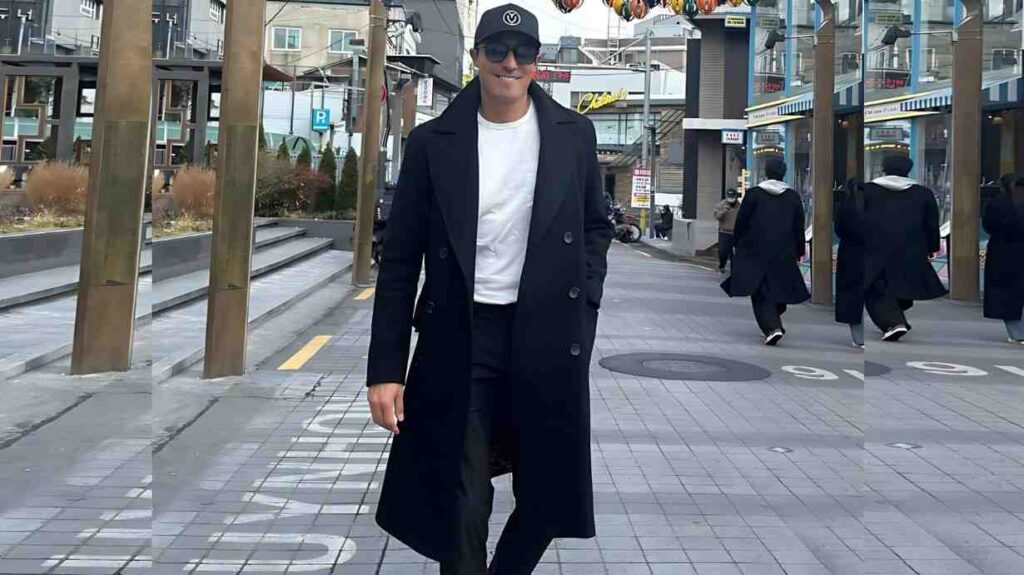 Daniel Henney in a long black coat, white tee, cap and sunglasses walks along a modern city street with glass storefronts.