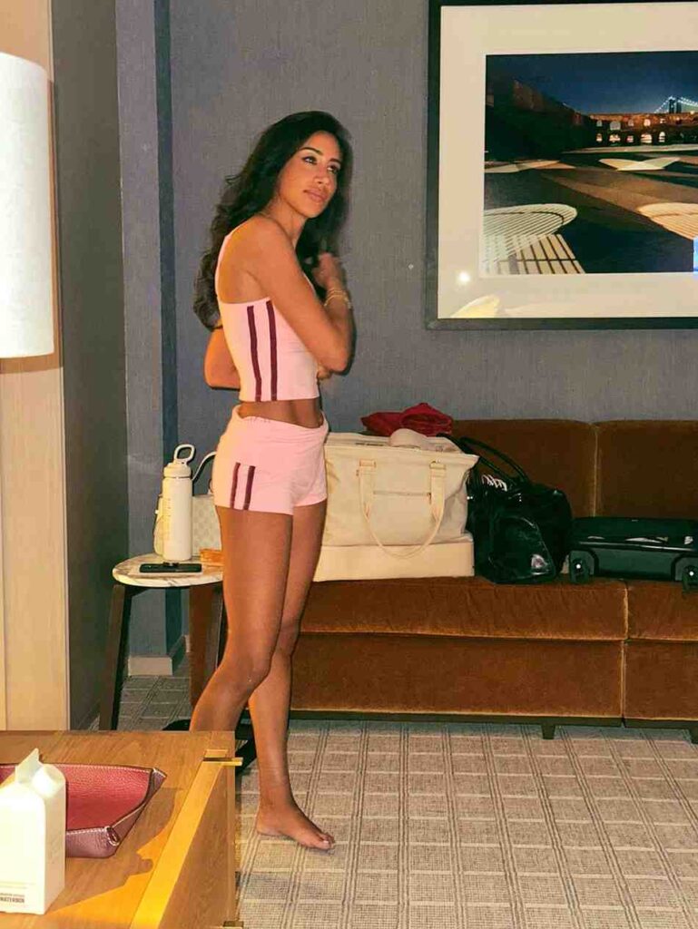 Danielle Jonas in pink sportswear stands with arms crossed in a modern living room, beside a beige sofa and a white water bottle.