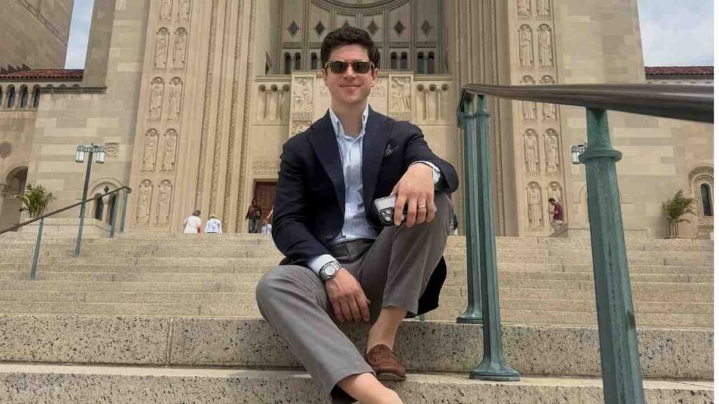 David Henrie in blazer and sunglasses sits on stone steps before an ornate, carved cathedral, holding a phone near a teal railing.