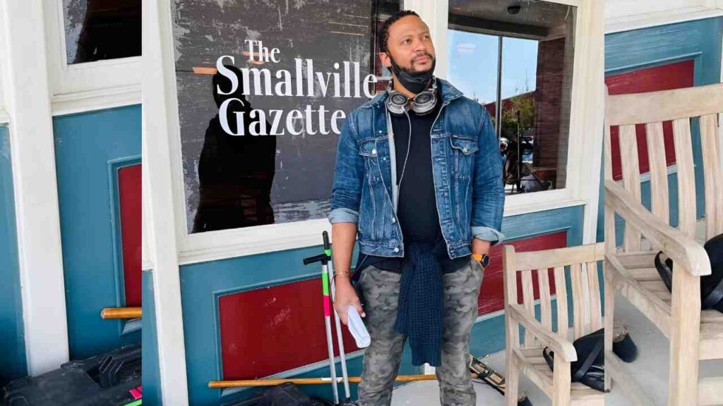 David Ramsey in a denim jacket and camo pants stands outside with 'The Smallville Gazette' sign, wearing headphones around his neck.