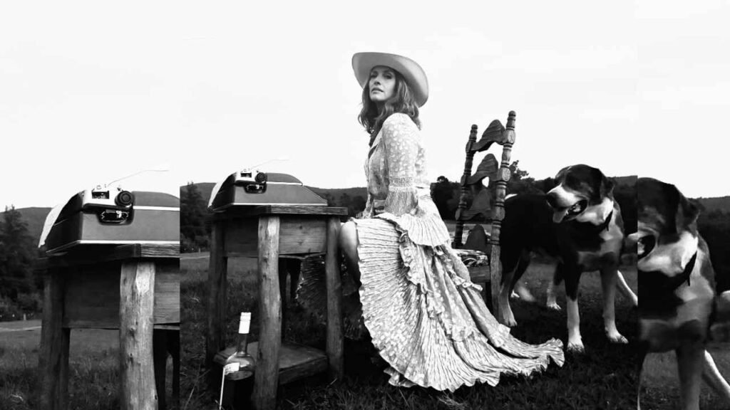 Dawn Olivieri in a wide-brim hat and flowing lace dress sits beside rustic grills, while two dogs stand nearby.