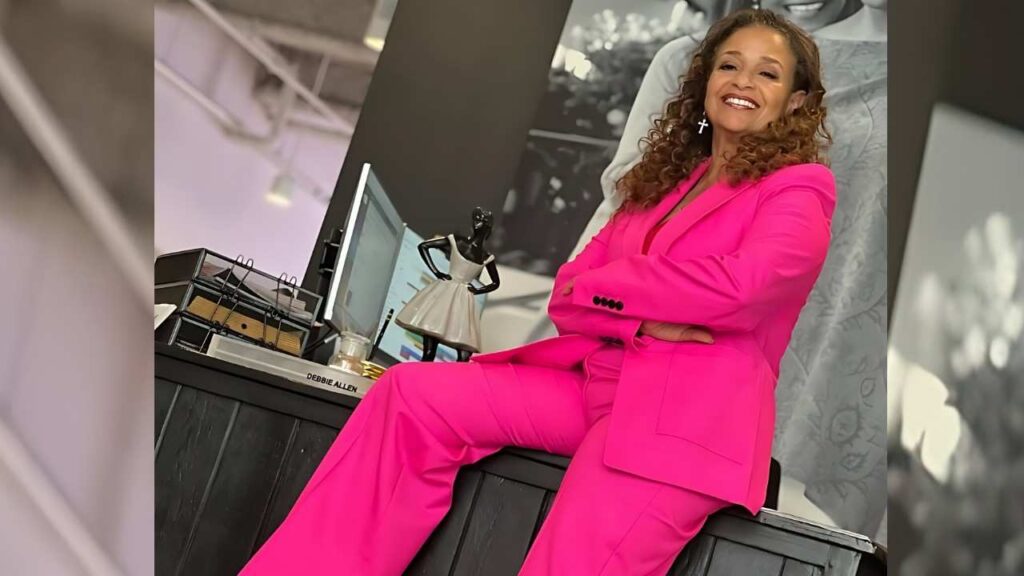 Debbie Allen in a bright pink pantsuit sits on a dark desk ledge in a modern office, arms crossed, curly hair, cross earrings, desk accessories nearby.