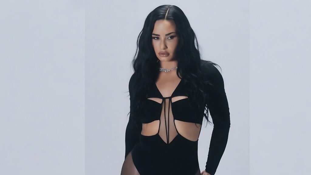 Demi Lovato with long black wavy hair in a black cutout bodysuit, sparkly necklace, posing against a light gray studio backdrop.