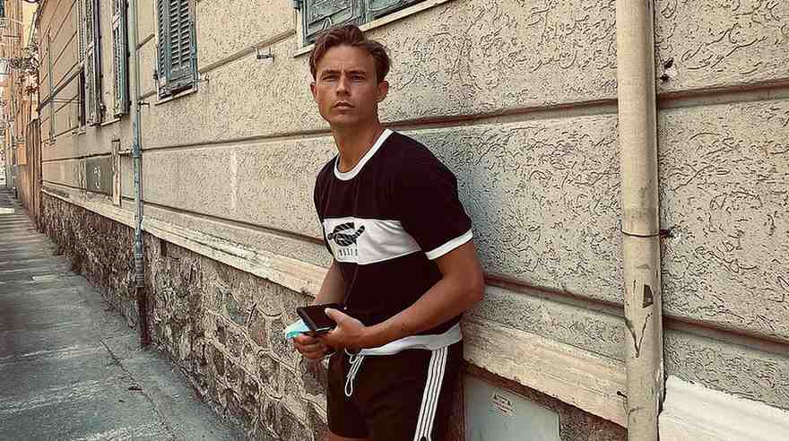 Dennis Mojen leaning against a beige wall in a narrow alley, wearing a black and white athletic outfit, holding a phone.