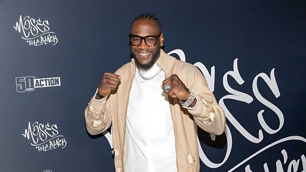 Deontay Wilder with braided hair and glasses poses fists up on a blue backdrop, wearing a beige ribbed jacket over white shirt.