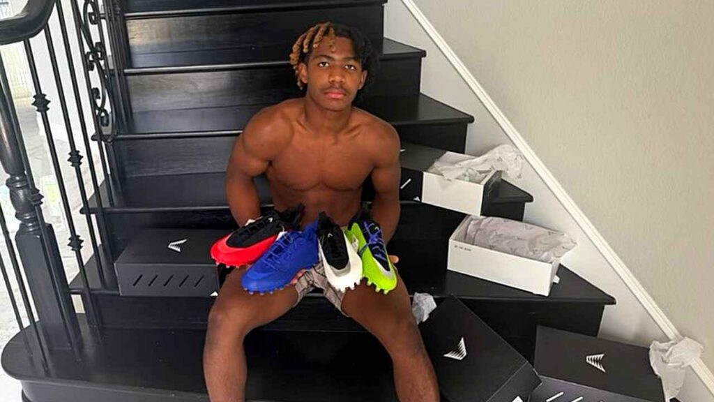 Dez Bryant with blond-tipped dreadlocks sits on black stairs, cradling five soccer cleats over his lap amid boxes.