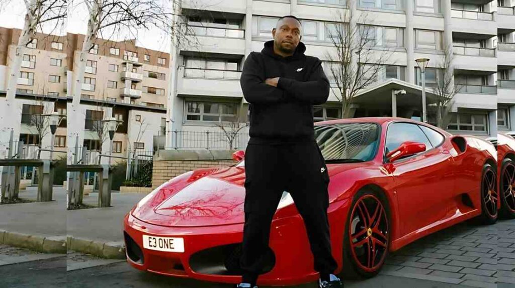 Dizzee Rascal in black hoodie and joggers stands with arms crossed beside a bright red Ferrari in a modern residential courtyard.