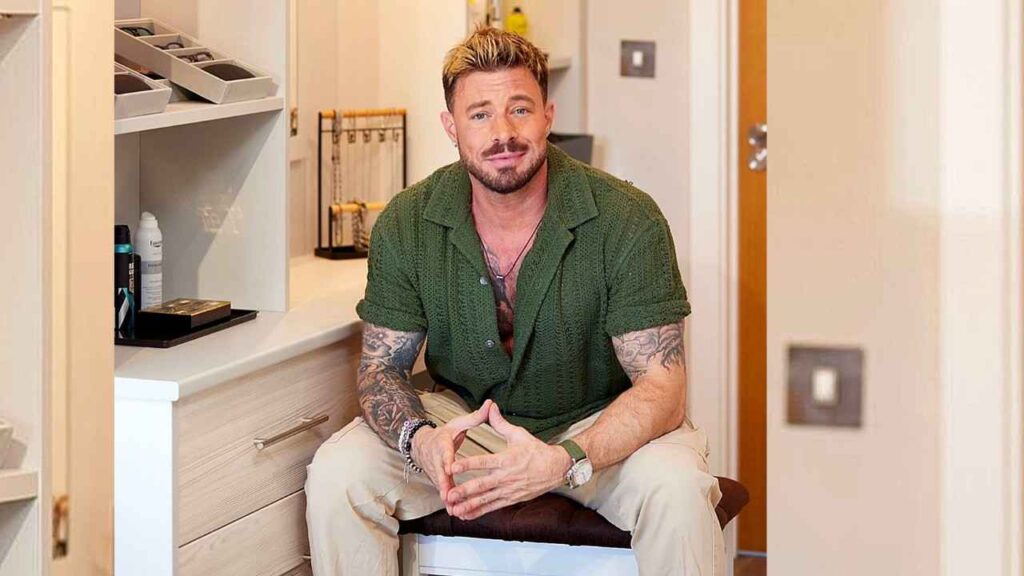 Duncan James with blond hair and stubble sits in a beige room, wearing a green shirt and khaki pants, hands clasped.