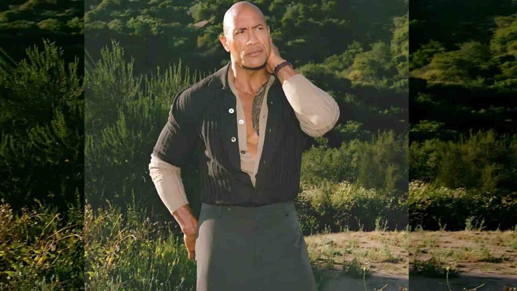 Dwayne Johnson wearing a dark open shirt over a light shirt and a long gray skirt; hand on his neck, the other on his hip, with green bushes behind.
