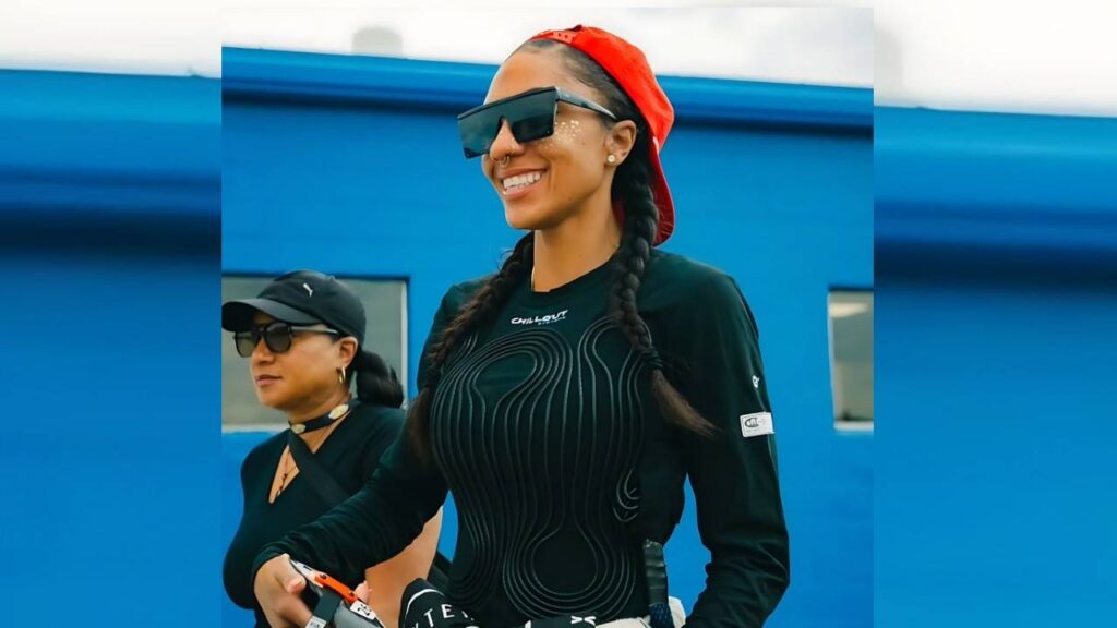 Dystany Spurlock with braids, black top, red cap, and oversized sunglasses by a blue wall; another woman follows behind.