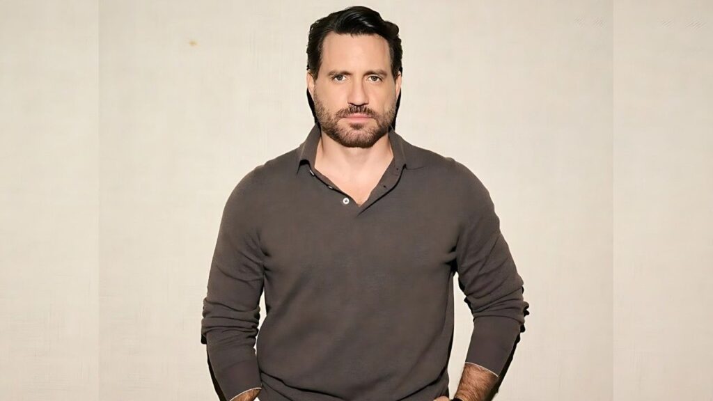 Edgar Ramírez with stubble in a gray long-sleeve polo stands against a pale beige studio backdrop, looking at the camera.
