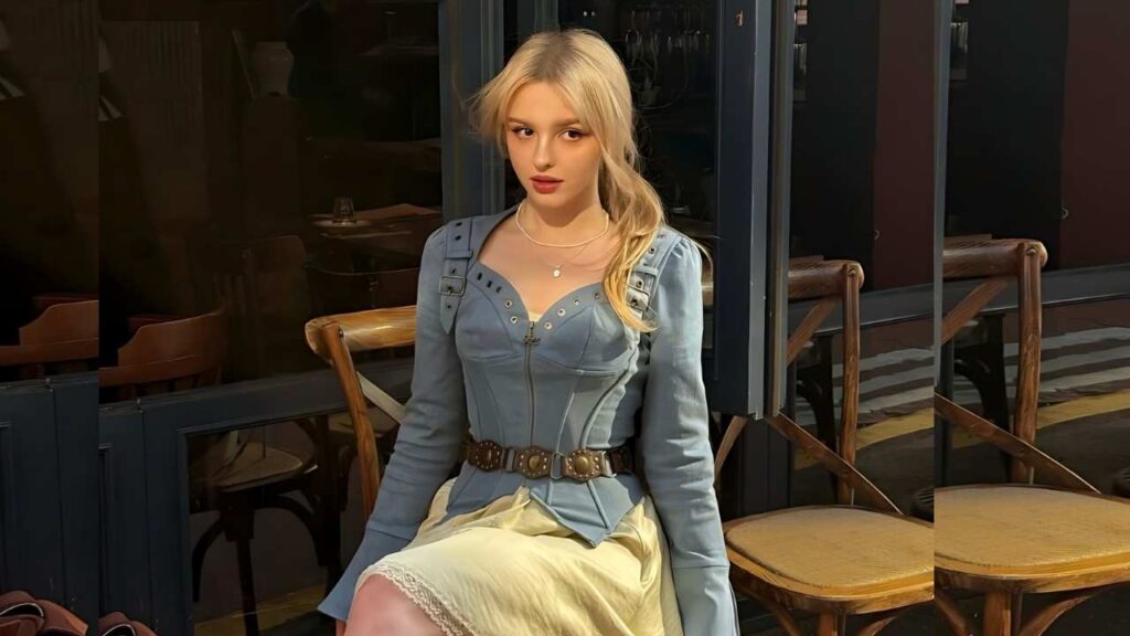 Elina Karimova sits outside a cafe in a blue corset-style top with a belt and pale yellow skirt, beside wooden chairs.