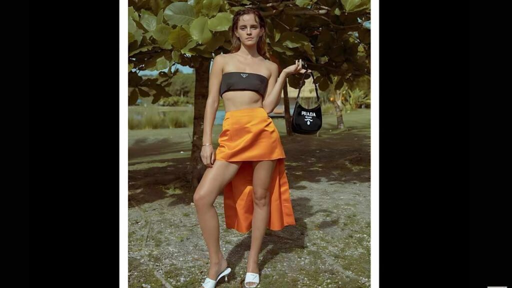 Emma Watson in a black strapless top and orange high-low skirt poses outdoors, holding a Prada bag near a lake.
