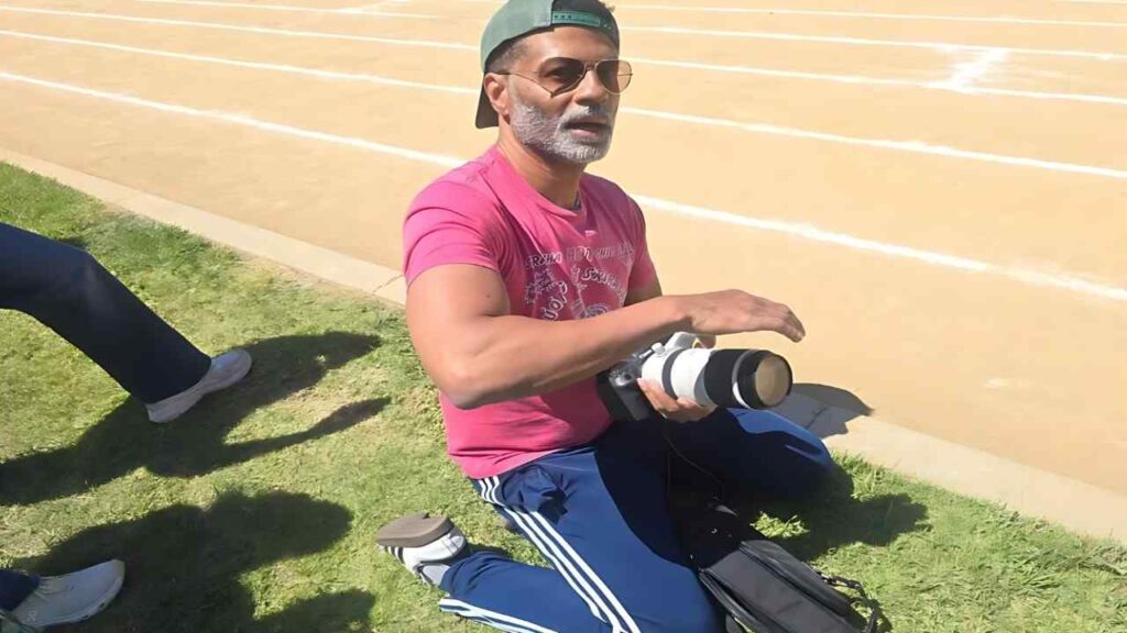 Eric Benét on grass beside a running track, wearing pink shirt, blue pants, sunglasses, a backward green cap, holding a telephoto lens.