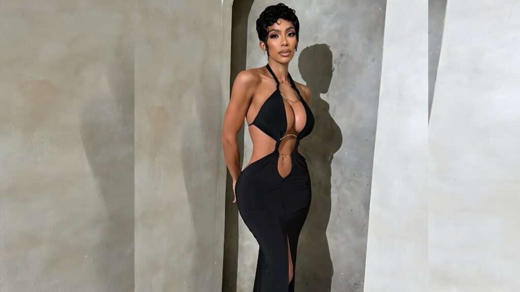 Erica Mena with short curly hair in a black cut-out halter jumpsuit poses against concrete wall, rings connecting fabric.