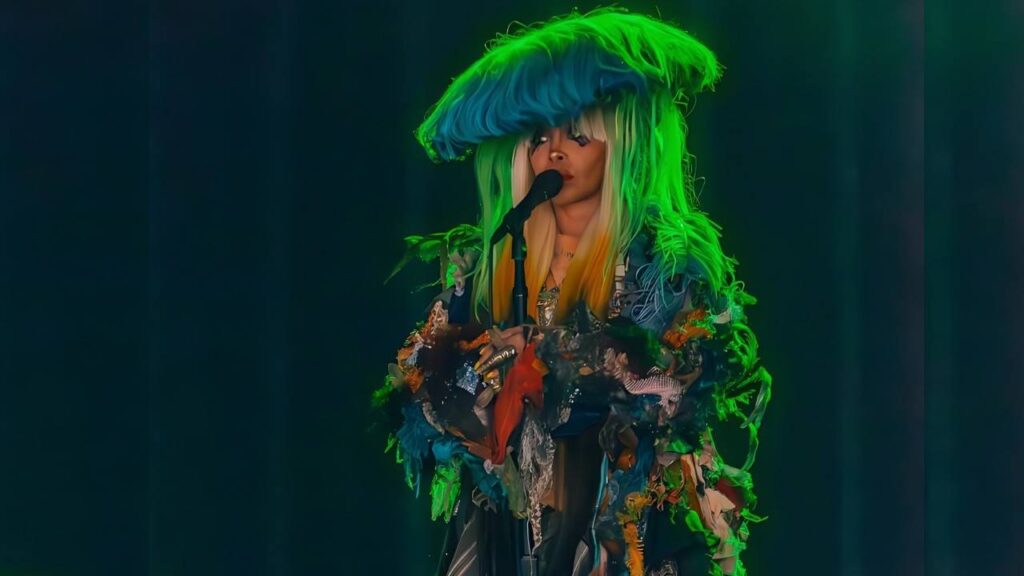Erykah Badu wearing a towering blue-green wig and patchwork costume sings into a microphone under green stage lights.
