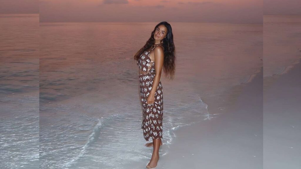 Eryn Krouse with long wavy hair stands barefoot on a sunset beach in a brown midi dress with a print, smiling at camera.