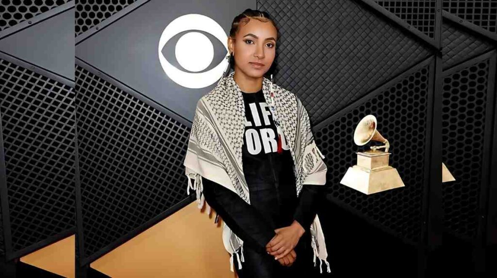 Esperanza Spalding in a shawl over black, standing before a dark backdrop with logo; a gramophone trophy sits to her right.