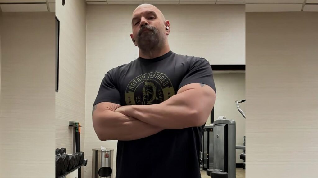 Ethan Suplee with a goatee stands arms crossed in a gym, wearing a dark graphic tee; weight machines visible behind him.