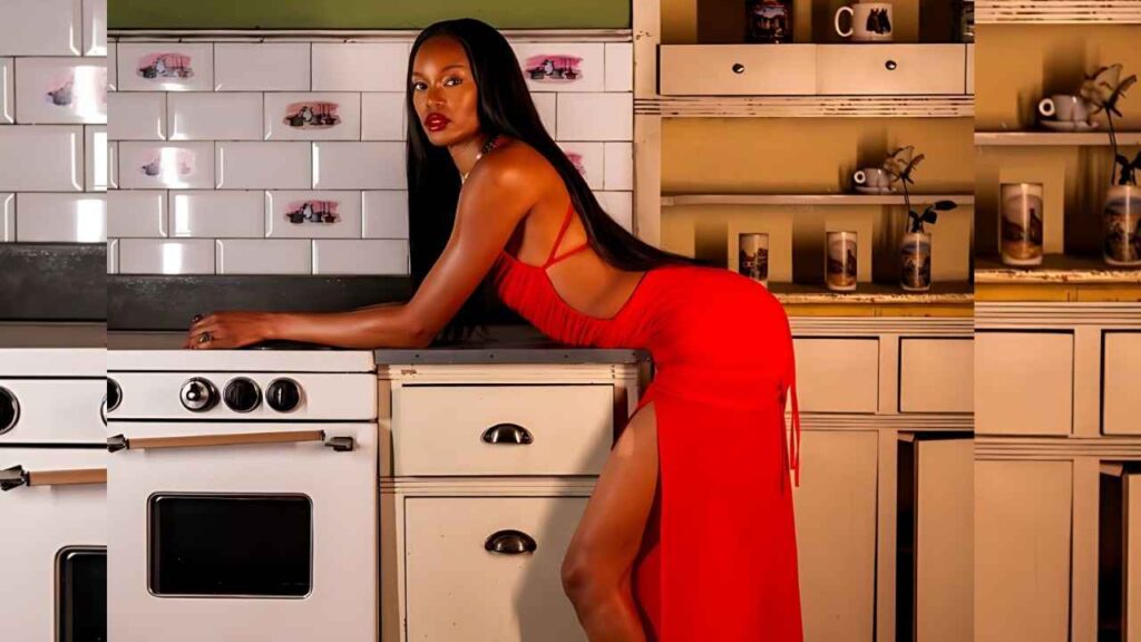 Eugena Washington in a red dress with a high slit leans on a vintage white stove in a retro kitchen, long black hair.