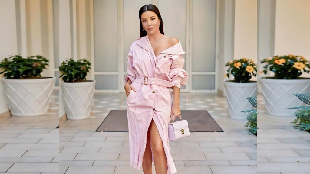 Eva Longoria in a pale pink belted trench standing in a light courtyard with white planters, holding a pink handbag.