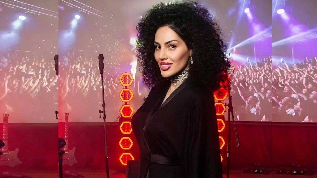 Eva Rivas with curly black hair in a black dress stands on a concert stage, red lights and an audience projection.