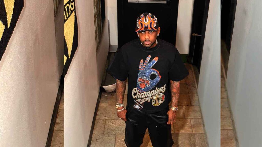 Fabolous in a hallway wearing a flame-pattern cap and a black oversized tee with a blue peace-sign and Champions graphic, black pants and bracelets.