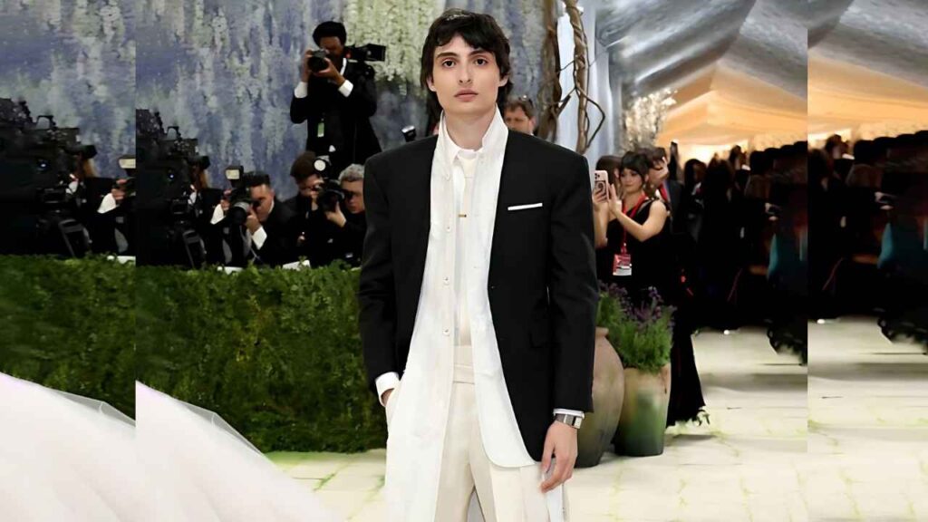 Finn Wolfhard in a black blazer and white shirt with cream trousers poses as paparazzi photograph him; green hedges behind.