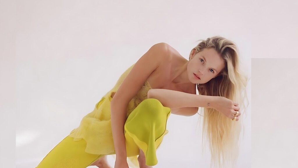 Gabriella Wilde in a bright yellow outfit crouches, leaning forward with long hair over her arm, against a pale studio backdrop.