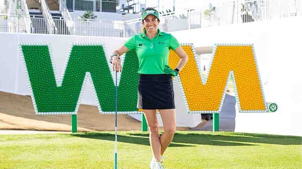 Gerina Piller in a green polo and black skirt stands on a golf course, resting a putter beside a large green-and-yellow sign.