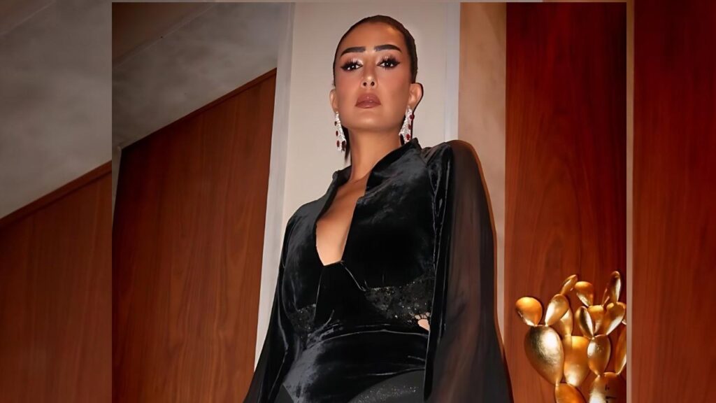 Ghada Abdel Razek in a black velvet dress with sheer sleeves poses against wood walls bold makeup and red chandelier earrings.