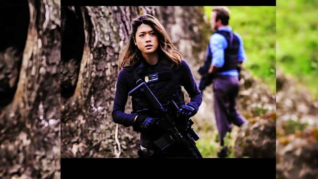 Grace Park tactical officer in black vest holding rifle in forest, teammate in background.