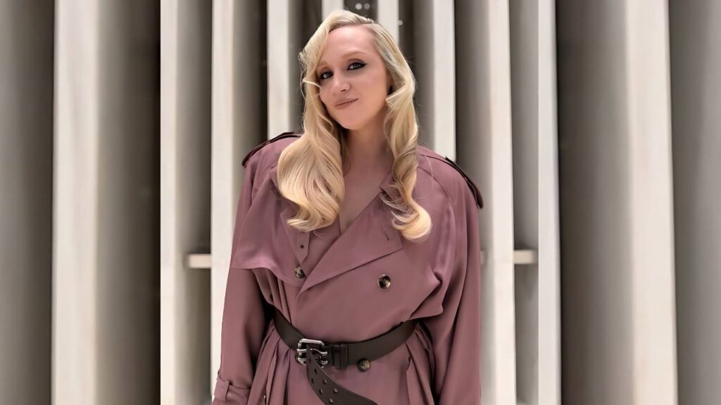 Gwendoline Christie in a mauve trench with a wide black studded belt stands before beige panels, curled hair and a subtle smile.
