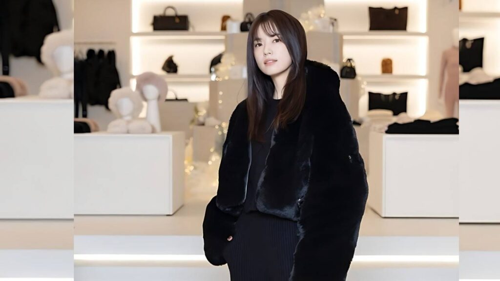 Han Hyo-joo with long dark hair in a black fur coat poses inside a bright boutique, shelves lined with handbags behind her.