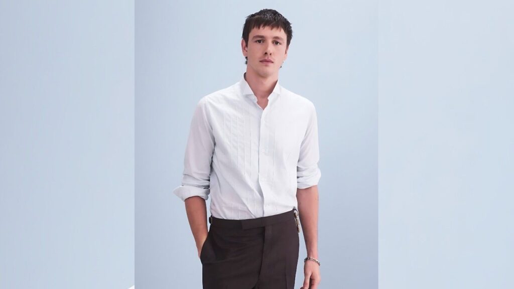 Harris Dickinson in white button-up shirt with rolled sleeves stands against light blue backdrop, hands in pockets, expression.