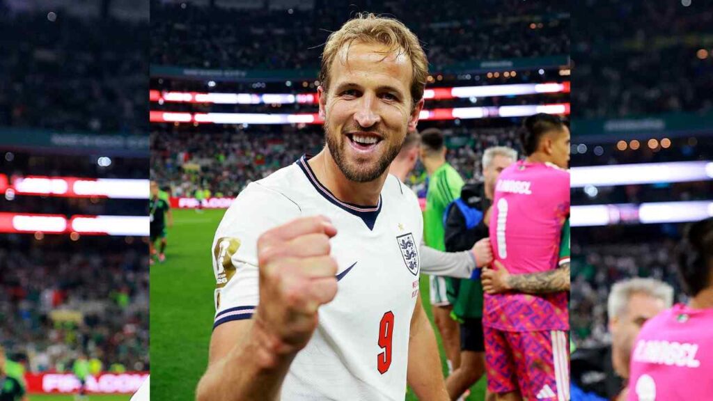 Harry Kane in white kit with number 9, fist raised toward the camera on a stadium packed with fans.