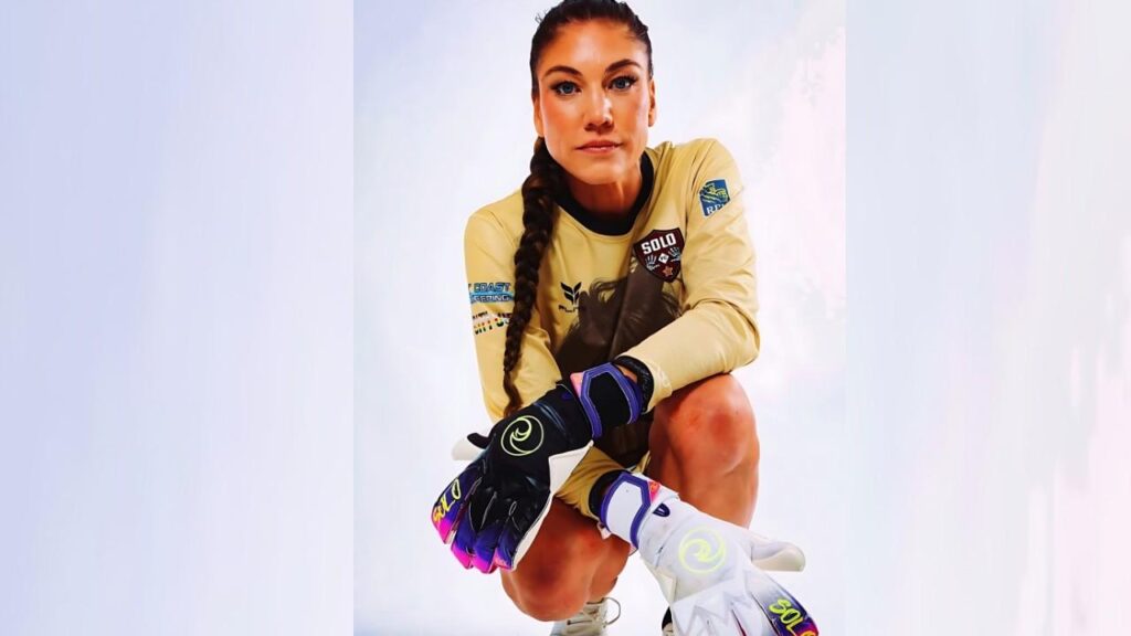 Hope Solo in a tan jersey with patches, kneels forward with braided hair and colorful gloves, intense gaze.