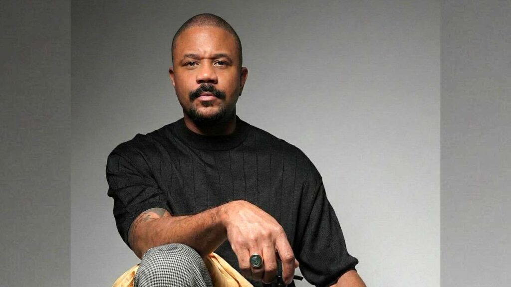 Hosea Chanchez with a trimmed beard in a black knit shirt, seated against a gray gradient, wearing a green ring.