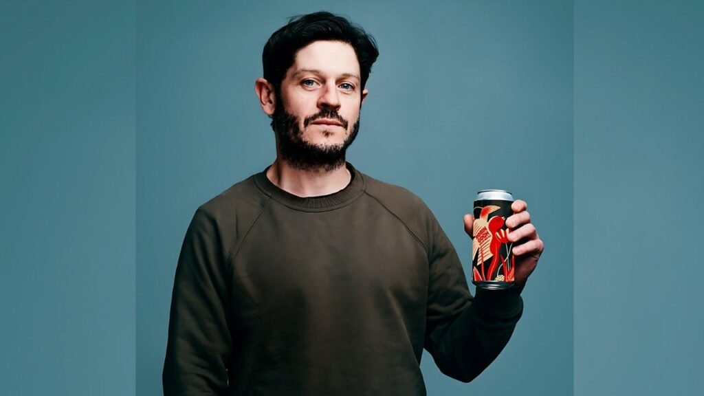 Iwan Rheon with dark hair and beard in an olive sweatshirt, holding a colorful can, posed against a teal backdrop with neutral gaze.