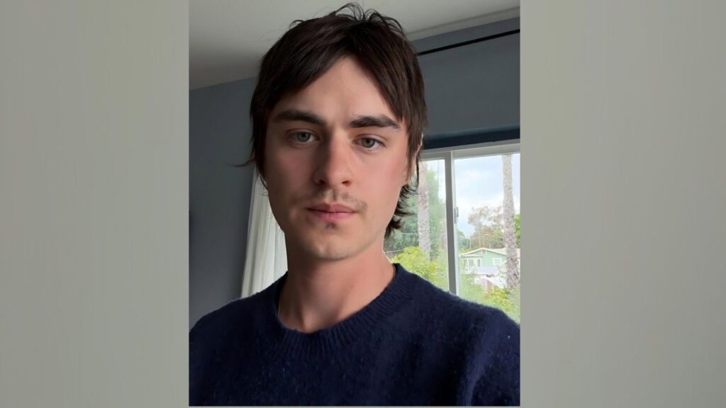 Jaeden Martell with dark hair and light stubble, wearing a navy knit sweater, indoors by a window with greenery outside.